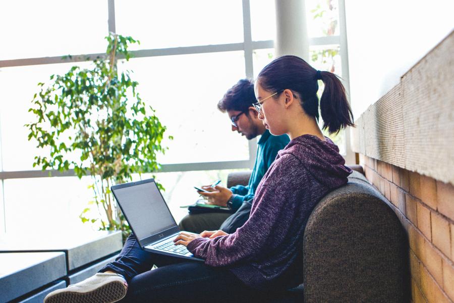 Two students sitting together. One is on their laptop and the other is checking his phone.
