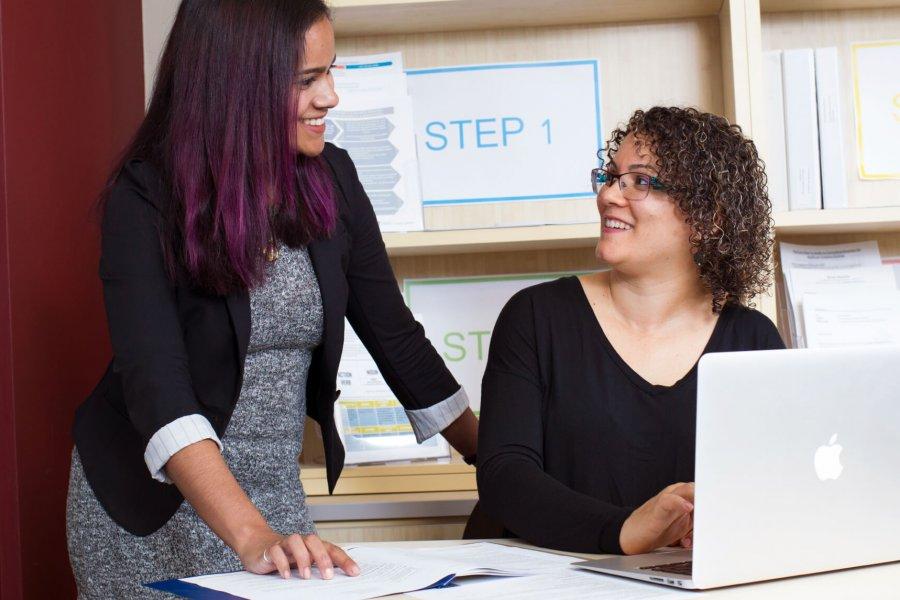 Two co-op students, one standing and one seated at a desk, review information in a notebook and on a laptop.