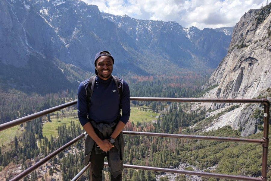 A student wearing a backpack stands at a lookout in the mountains of British Columbia, a beautiful valley behind him.