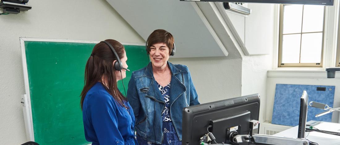 Two members of the Faculty of Social work work together in an office wearing headsets and standing in front of a computer work station.