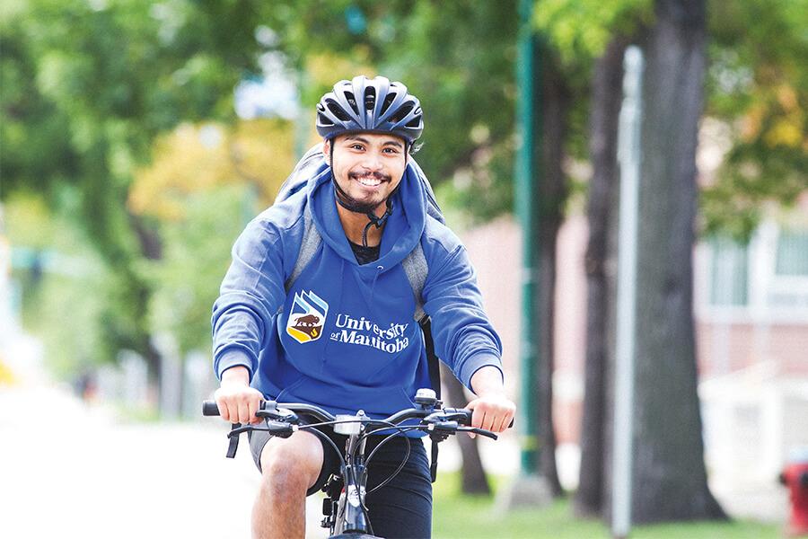 A smiling student riding a bike, wearing a helmet and U M hoodie.
