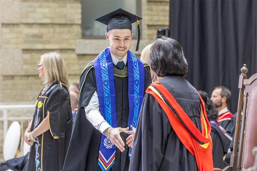 A student wearing a cap and gown with an indigenous sash shaking hands with a professor.
