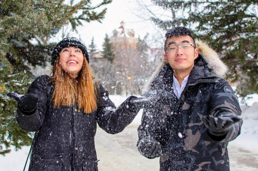 A student mentee and mentor stand together outside on a winter day smiling and playfully tossing snow in the air. 