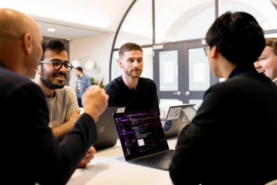 Group of people sitting around a table with laptops, engaged in a collaborative discussion in a bright indoor space.