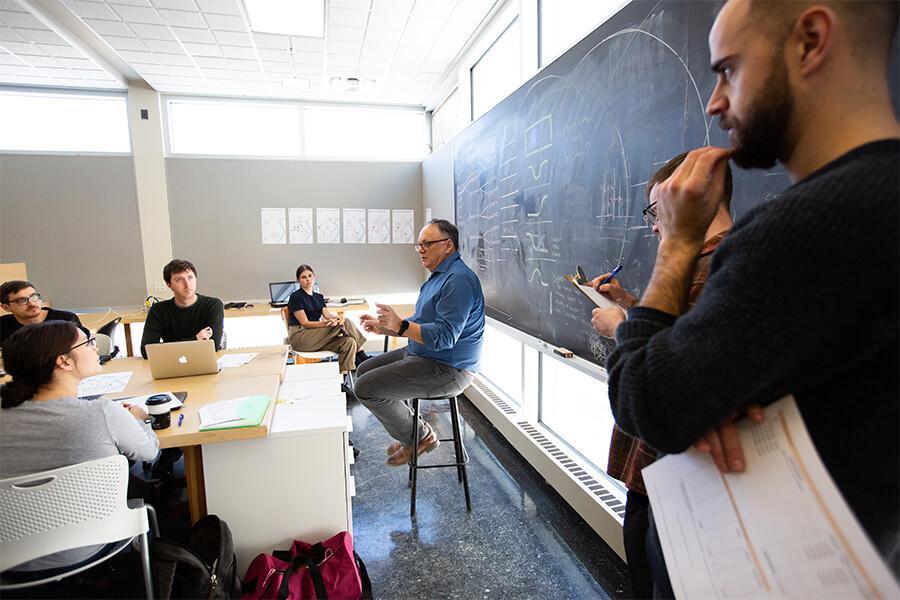 An image of a professor lecturing their engaged students inside of a classroom on a sunny day