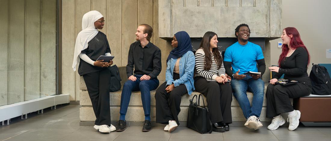 Six students are gathered casually in a bright indoor space with concrete walls and large windows. One student wearing a white headscarf and black outfit stands holding a tablet and talking to the group. Five others sit on a low concrete bench, engaged and smiling; they include people with varied appearances and clothing styles, from business casual to relaxed jeans and sneakers. Some hold notebooks or tablets, and backpacks are nearby, suggesting a collaborative or social study moment.