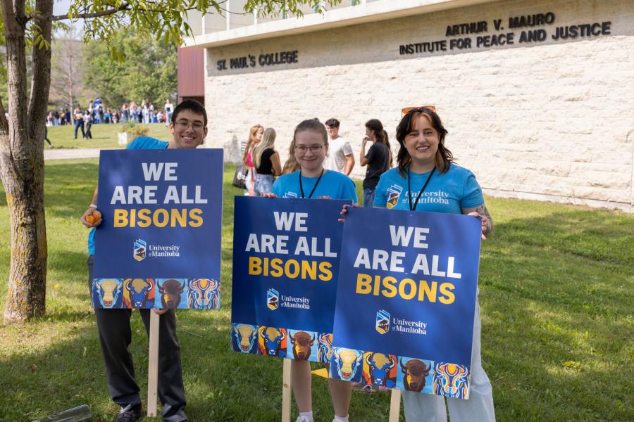 three um orientation volunteers stand with smiling faces in front of st paul's college with "We Are All Bisons" signs in hand.