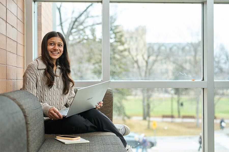 A student sits on a modern indoor bench by large windows, smiling while working on a laptop. A notebook and pen rest beside her, and outside the window are bare trees and campus buildings.