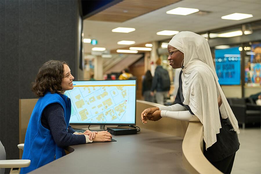 A student wearing a light headscarf speaks with a campus staff member at an information desk. The staff member smiles and gestures while a large computer monitor displays a campus map behind them.