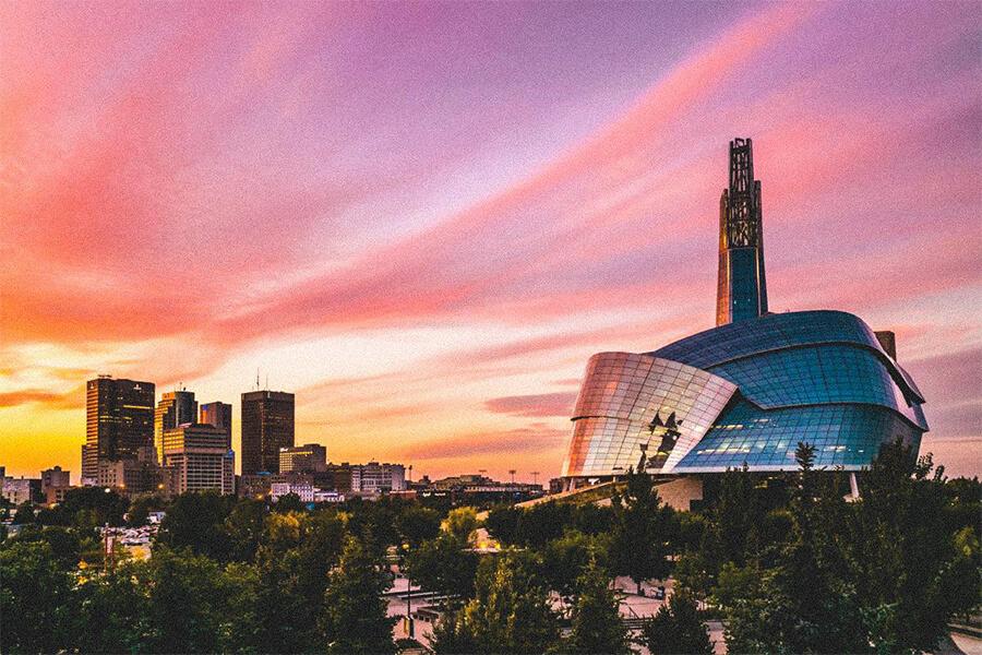 A vibrant sunset sky over downtown Winnipeg with the Canadian Museum for Human Rights in the foreground. Modern skyscrapers and tree-lined streets are visible under dramatic pink and orange clouds.