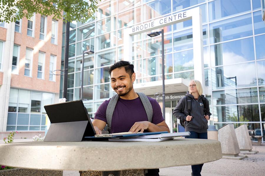 A student looking at a laptop on a cement picnic table outside the Brodie Centre.