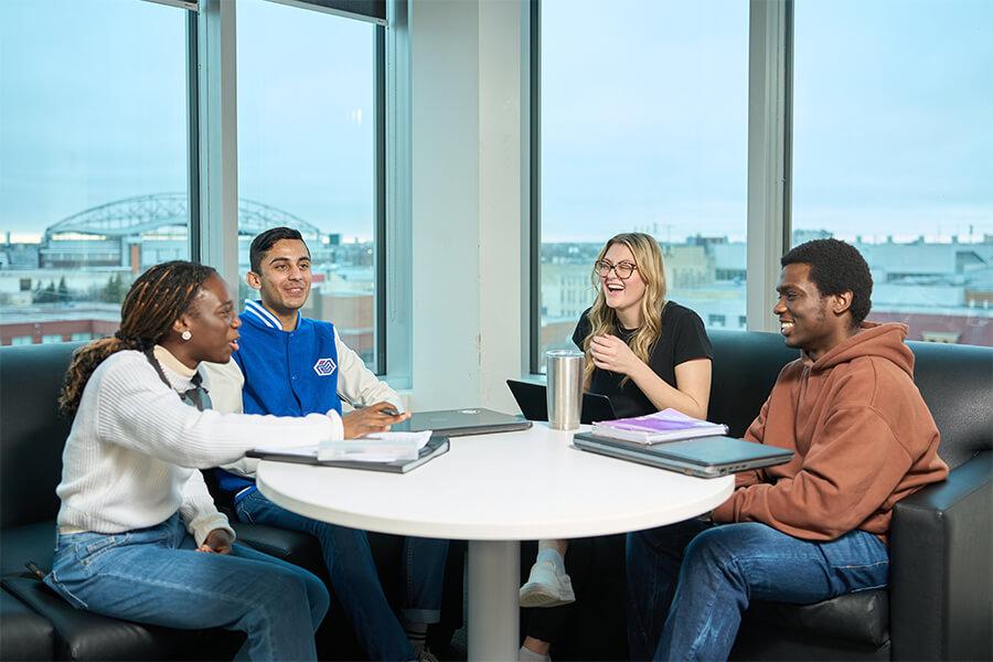 Four students sit around a round table in a bright lounge area with city views behind them. They’re smiling and chatting while working on laptops and notebooks, creating a friendly study environment.