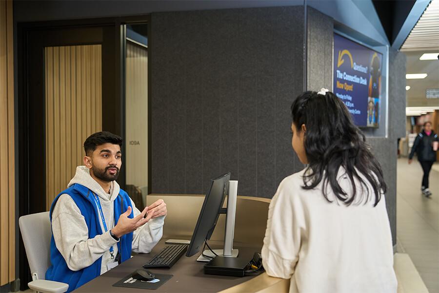 A student at an information desk speaks with a campus staff member in a blue vest. The staff member gestures while explaining something, and a sign reading “Questions? The Connect Desk Now Open!” is visible in the background.