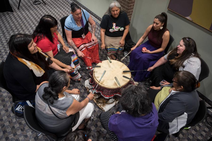 A group of women gathered in a circle beating a PowWow drum.