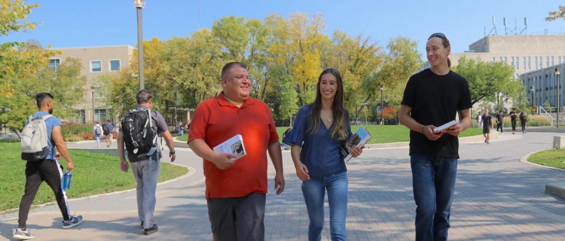 Three people walking and talking on the University of Manitoba campus, holding books and smiling on a sunny day.