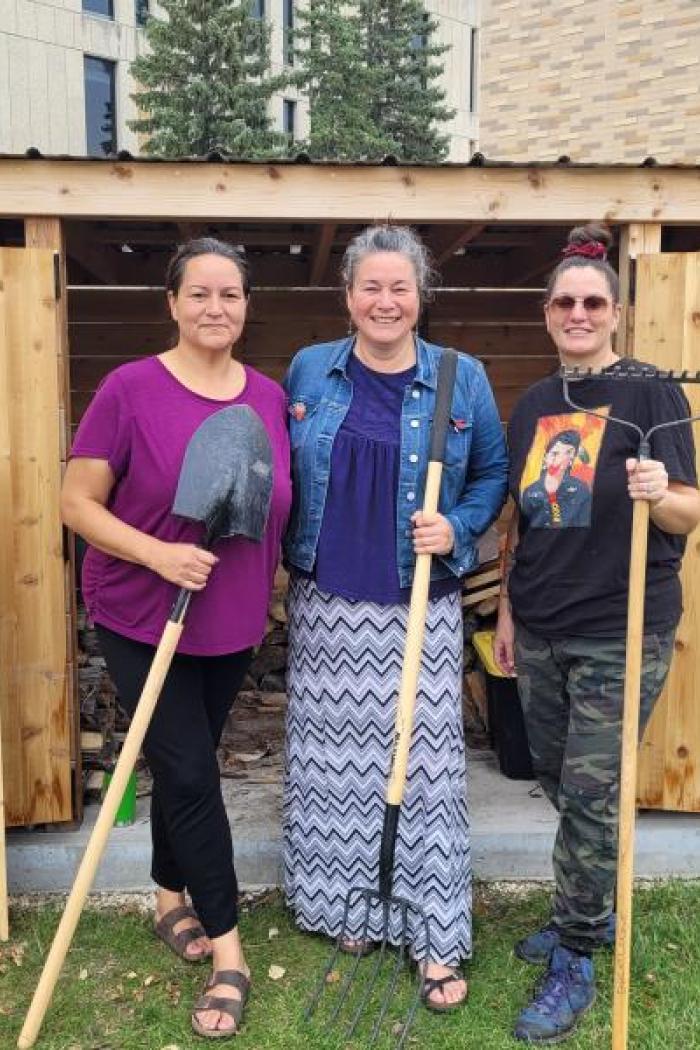 Christine Cyr, Nicki Ferland, and Vanessa Lillie standing together in front of the sweat lodge, holding gardening tools and smiling.