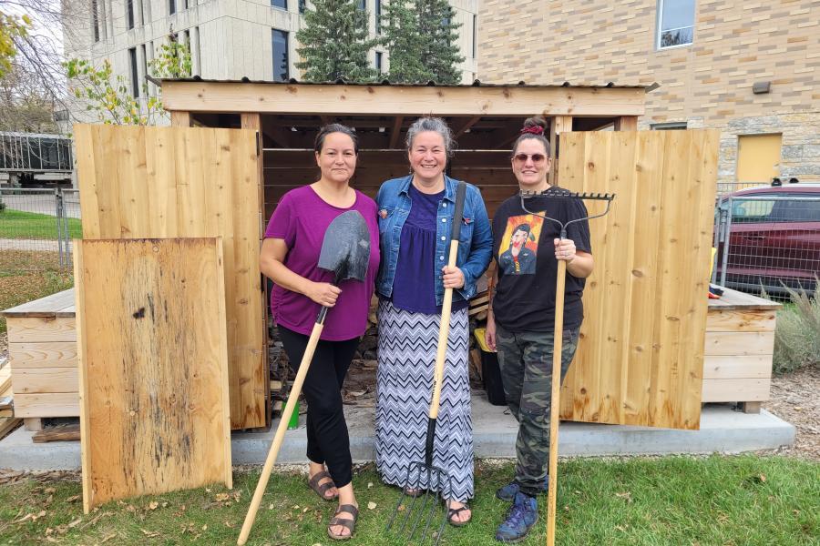 Christine Cyr, Nicki Ferland, and Vanessa Lillie standing together in front of the sweat lodge, holding gardening tools and smiling.
