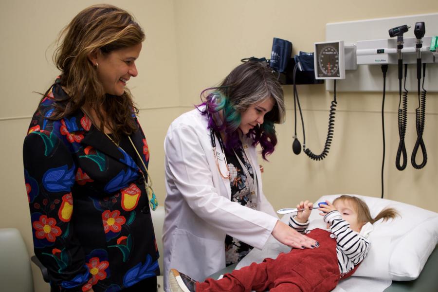 Two Ongomiizwin health service providers observe a smiling child to ensure she is healthy. 