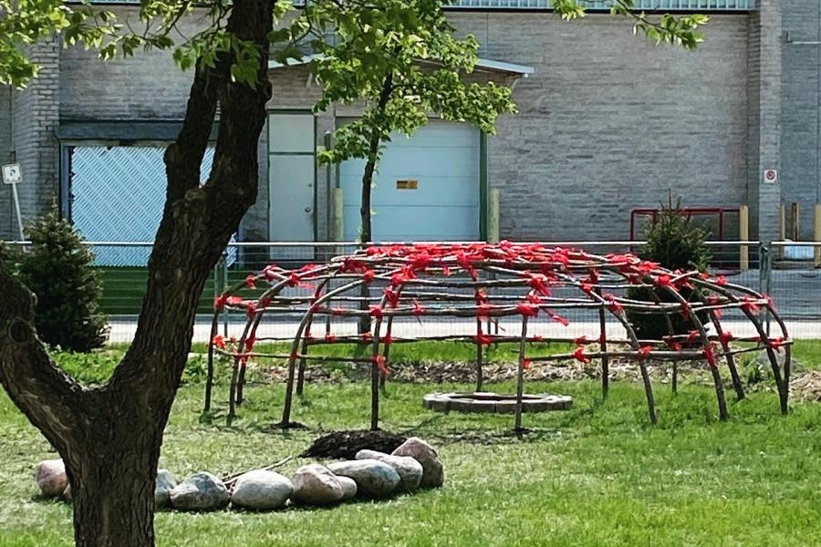 A sweat lodge tied with red ribbons on a grassy area with a tree in the foreground