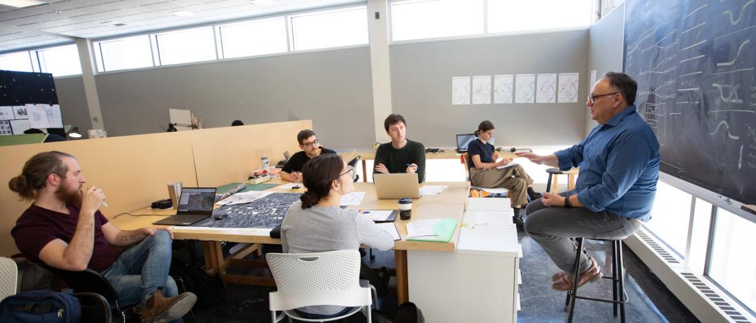 A professor sits at teh front of a classroom and speaks to a group of five students.