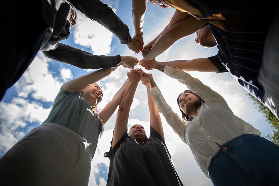looking ground up at a group of six people standing in a circle and holding their arms up, together.