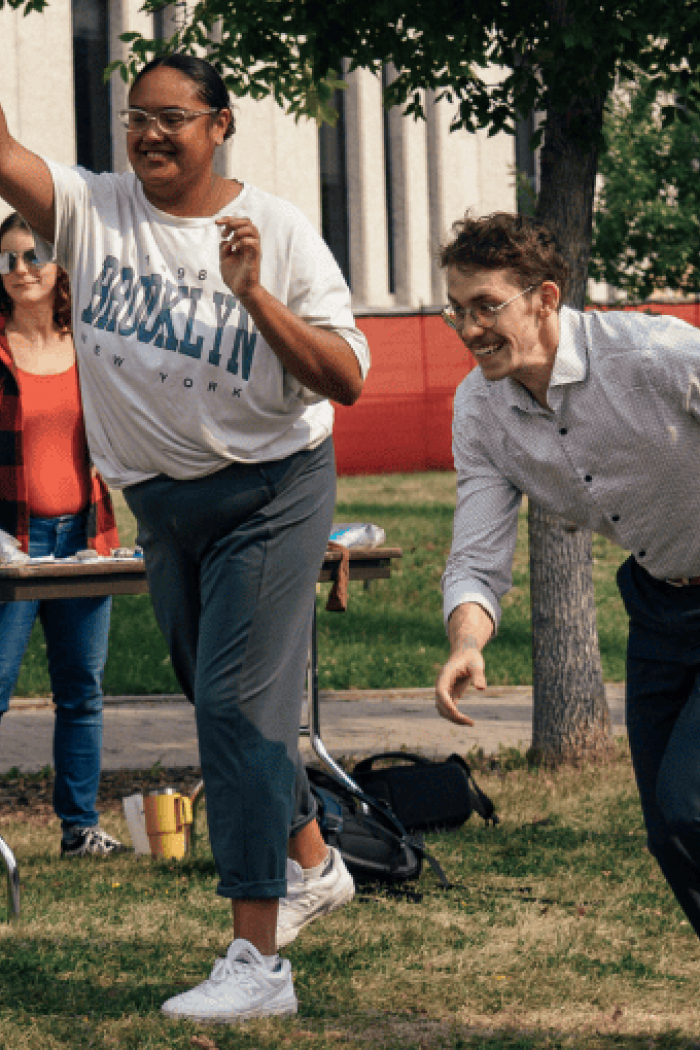 Two people play a lawn game outdoors during a campus event, tossing rings toward a target while others watch and help at a nearby table