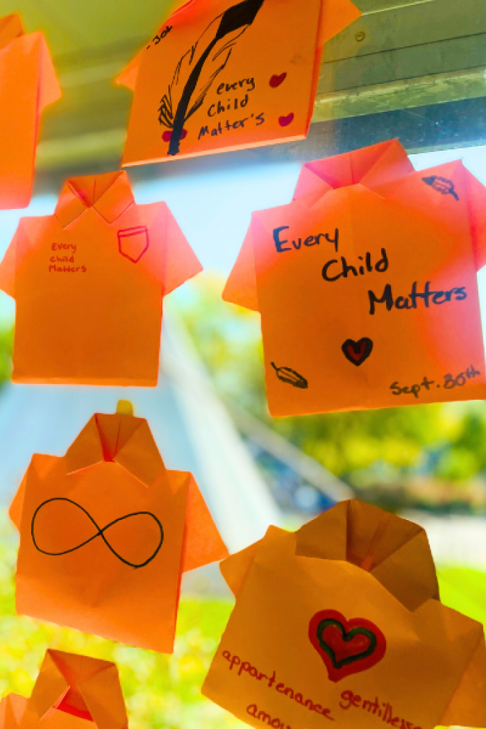 Orange paper shirts with handwritten “Every Child Matters” messages displayed on a sunny window, symbolizing support for residential school Survivors.