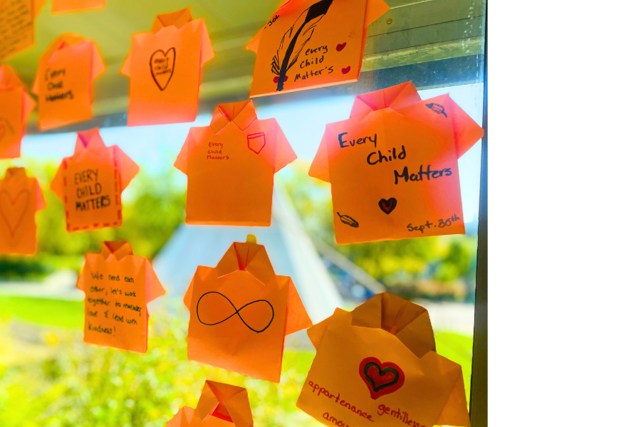 Orange paper shirts with handwritten “Every Child Matters” messages displayed on a sunny window, symbolizing support for residential school Survivors.