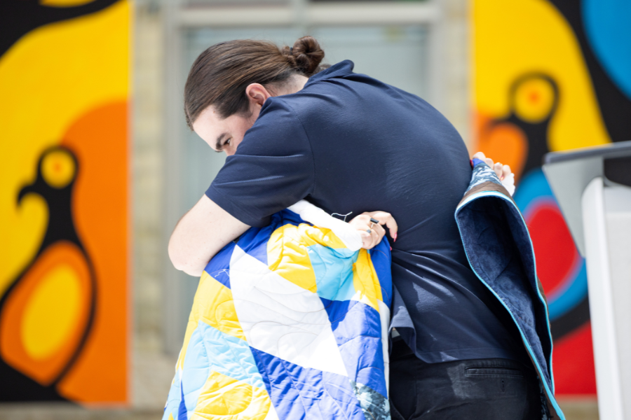 Young man wrapped in a star blanket hugs another person during the blanketing ceremony.