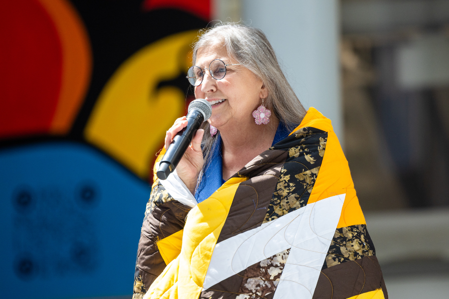 Woman wrapped in a star blanket speaks at the podium.