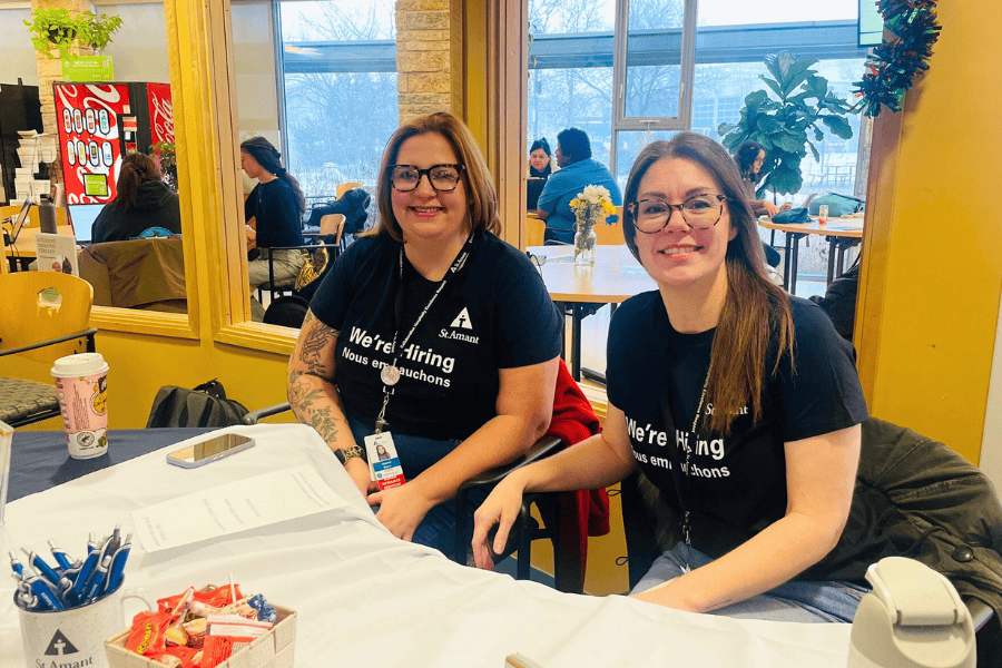 Two staff members wearing “We’re Hiring” shirts seated at a recruitment table with forms and pens in a campus common area.