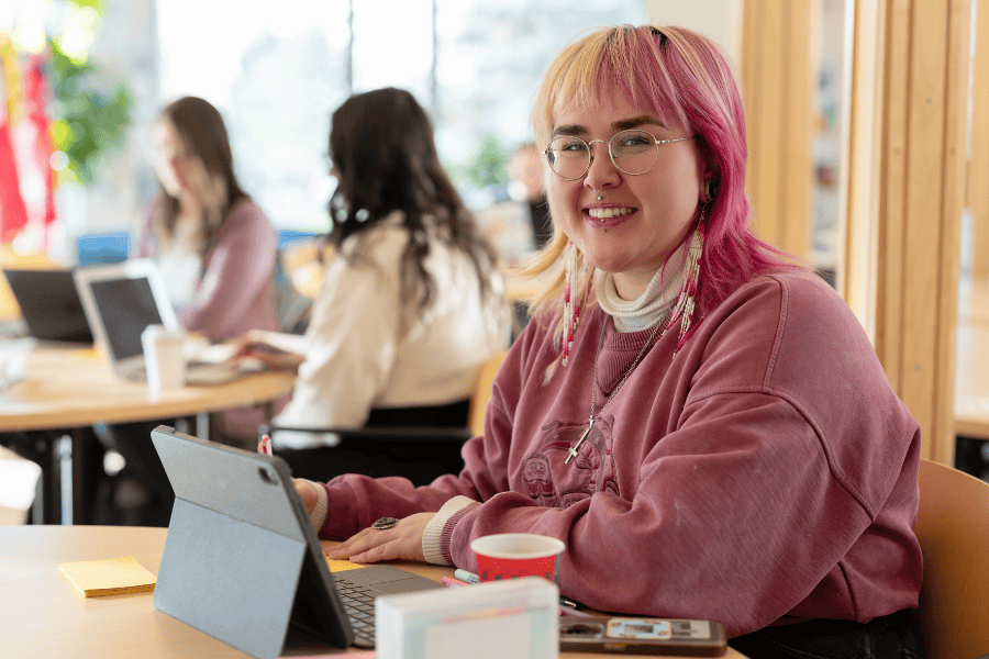 Student with pink hair and glasses smiling while using a tablet at a table in a bright study space, with other students working in the background.