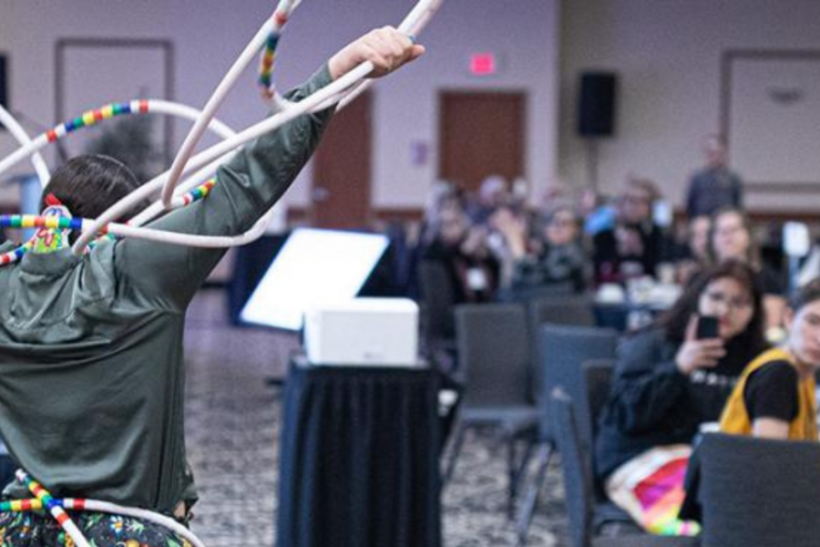 Performer presenting a hoop dance on stage at a community gathering, with audience members watching and clapping.