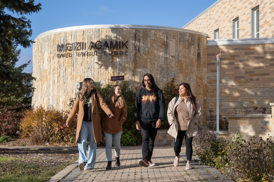Four Indigenous students walk and talk together outside Migizii Agamik – Bald Eagle Lodge at the University of Manitoba on a sunny day, surrounded by fall foliage.