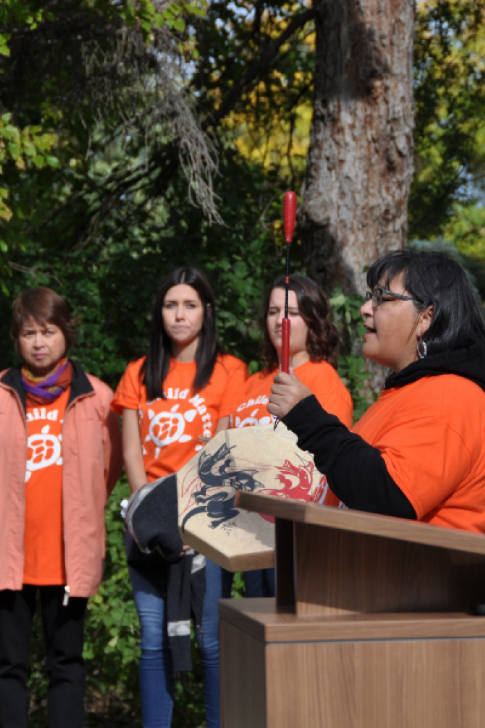 Woman speaking at podium holding a drum during Orange Shirt Day, with others in orange shirts listening outdoors.