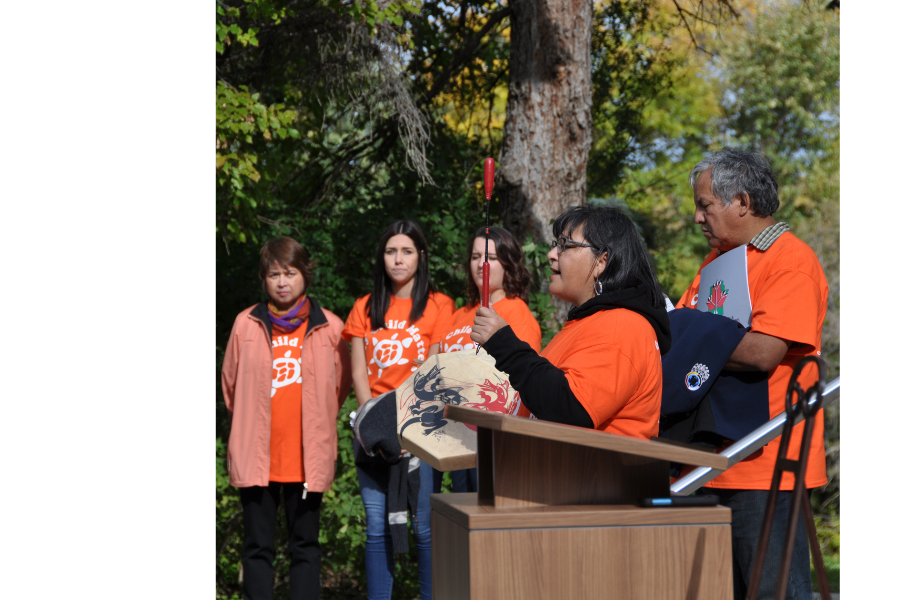 Woman speaking at podium holding a drum during Orange Shirt Day, with others in orange shirts listening outdoors.