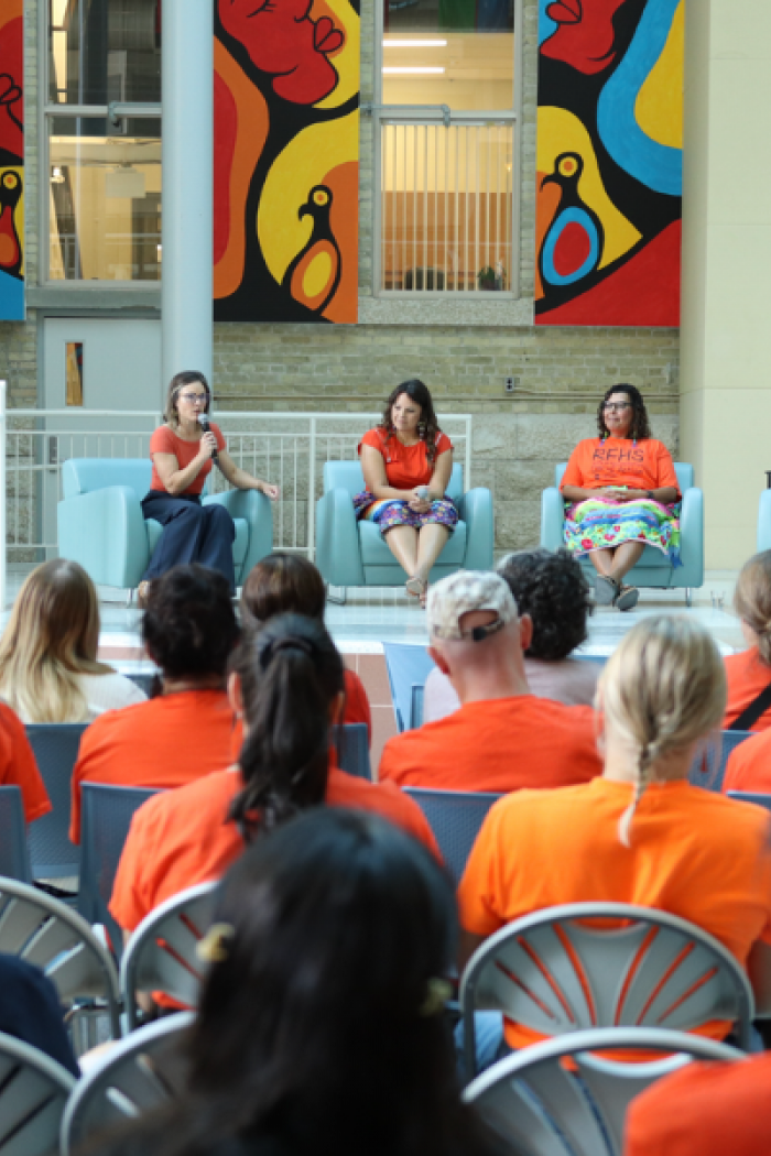 Three speakers sit on stage for a panel discussion, addressing an audience wearing orange shirts during an Orange Shirt Day event.