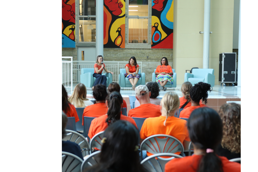 Three speakers sit on stage for a panel discussion, addressing an audience wearing orange shirts during an Orange Shirt Day event.