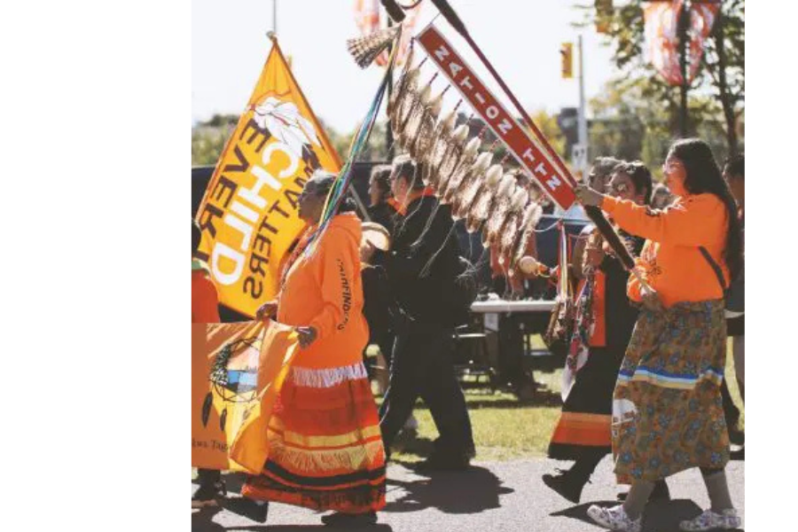 People wearing orange shirts and regalia participate in a Truth Walk, carrying flags and a staff with feathers.