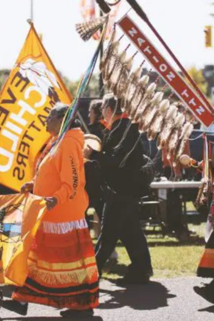 People wearing orange shirts and regalia participate in a Truth Walk, carrying flags and a staff with feathers.