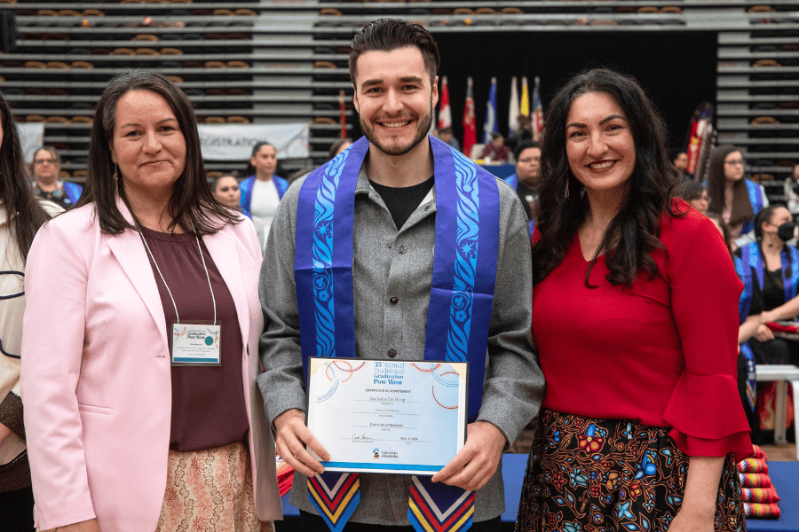 Graduate wearing a ceremonial stole holds a certificate, standing with two presenters at Grad Pow Wow.
