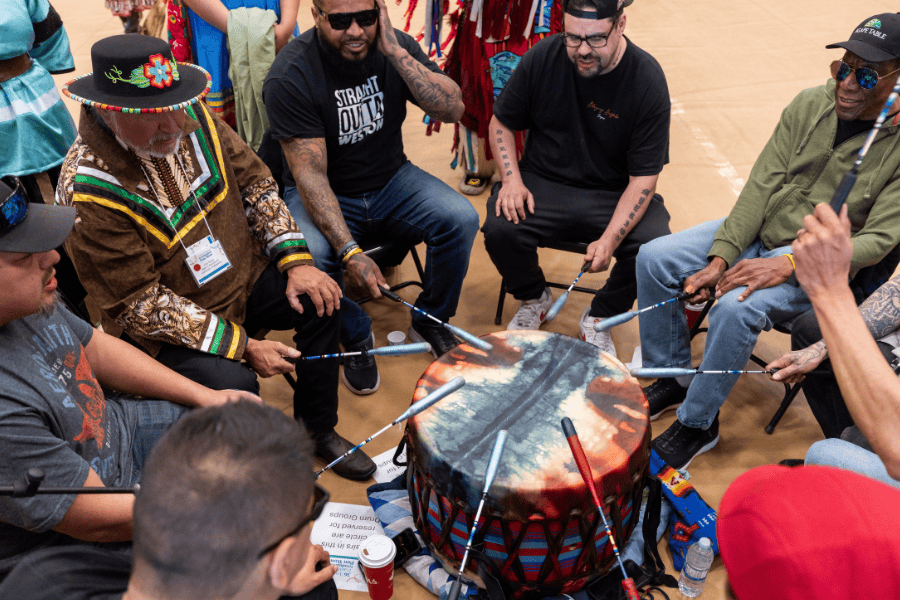 Drummers sit in a circle, playing a large Pow Wow drum together.