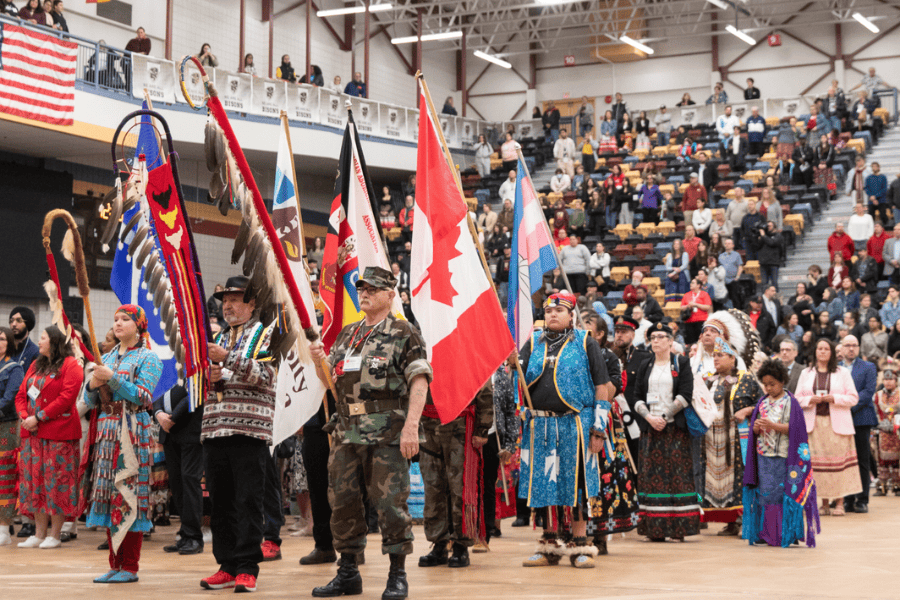 Indigenous participants in regalia carry flags during a Grand Entry at an indoor Pow Wow, with a large audience in the background.