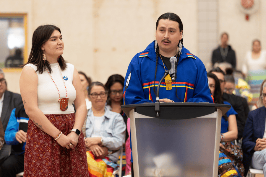 A student speaks at a podium during the Grad Pow Wow, with another participant standing beside him.