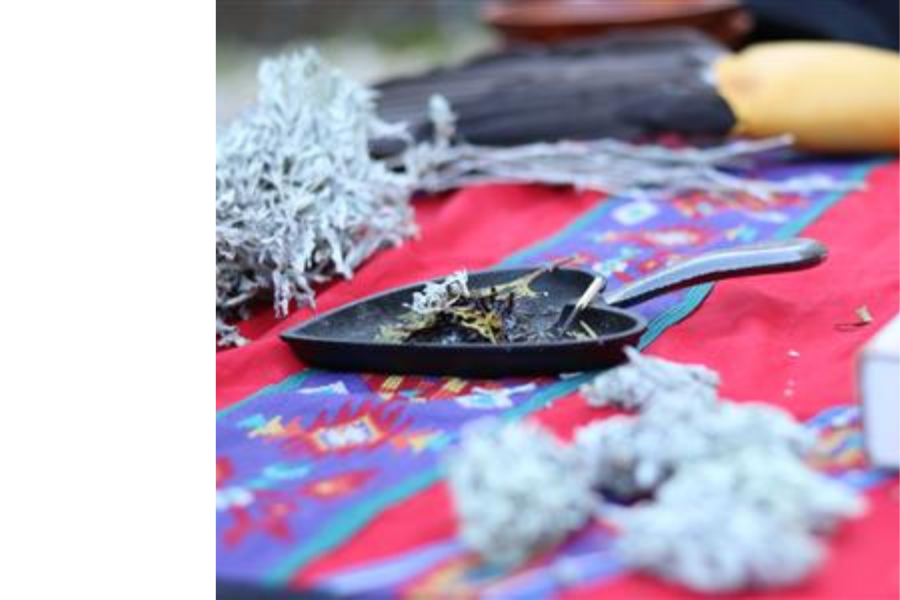 Close-up of smudging materials on a colorful cloth, including sage, a smudge pan with burning herbs, and other ceremonial items.
